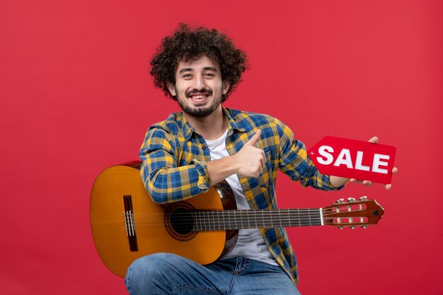 Vue de face jeune homme assis avec guitare sur mur rouge musique live applaudissements concert vente jouer