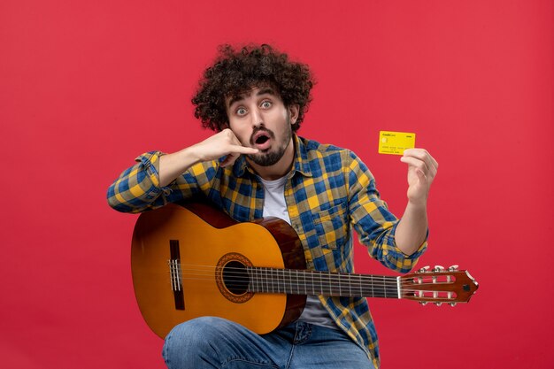 Vue de face jeune homme assis avec guitare tenant une carte bancaire sur le mur rouge concert de musique applaudissements couleur live