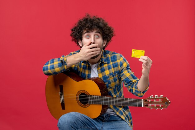 Vue de face jeune homme assis avec guitare tenant une carte bancaire sur le mur rouge concert de musique applaudissements musicien couleurs vente en direct