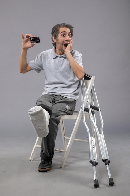 Vue de face jeune homme assis avec pied cassé et bandage tenant une carte bancaire sur un mur gris torsion homme accident douleur au pied jambes