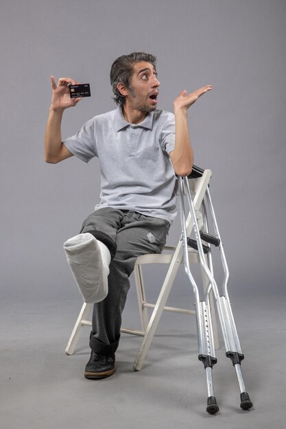 Vue de face jeune homme assis avec pied cassé et bandage tenant une carte bancaire sur le mur gris torsion homme douleur au pied accident