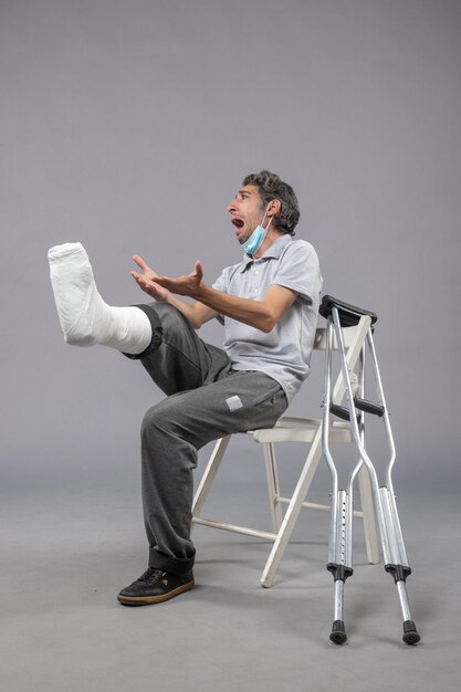 Vue de face jeune homme assis avec un pied cassé bandé criant de douleur sur le mur gris douleur à la jambe accident torsion pied mâle