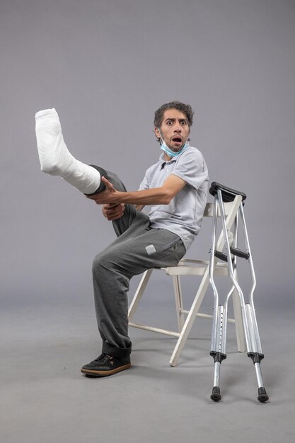 Vue de face jeune homme assis avec un pied cassé bandé sur un mur gris douleur à la jambe accident torsion pied mâle