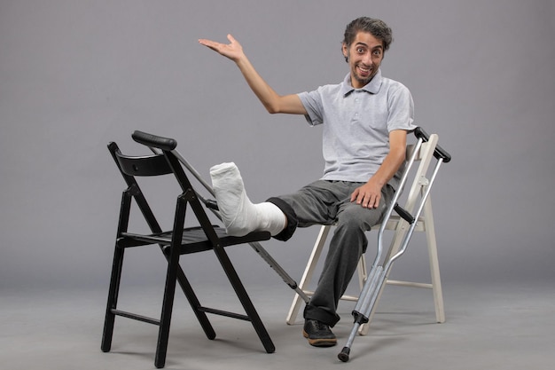 Vue de face jeune homme assis avec pied cassé et béquilles sur le mur gris accident cassé torsion pied douleur jambe