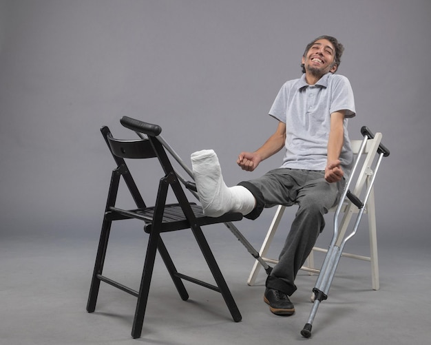 Photo gratuite vue de face jeune homme assis avec pied cassé et béquilles sur le mur gris accident torsion douleur à la jambe pied cassé