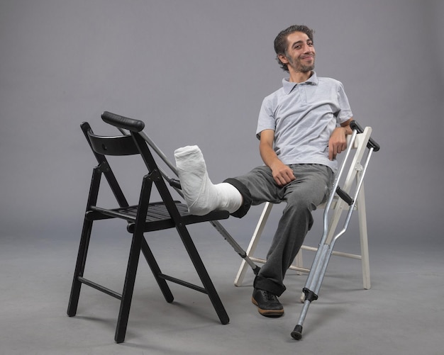 Vue de face jeune homme assis avec pied cassé et béquilles sur le mur gris accident torsion pied cassé douleur à la jambe