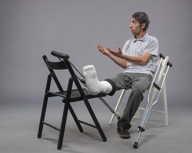 Photo gratuite vue de face jeune homme assis avec pied cassé et béquilles sur un mur gris douleur accident torsion jambes cassées