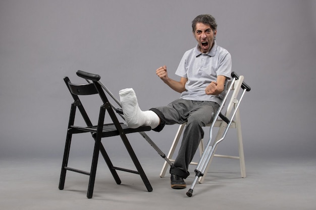 Vue de face jeune homme assis avec pied cassé et béquilles sur mur gris douleur au pied accident jambe cassée torsion