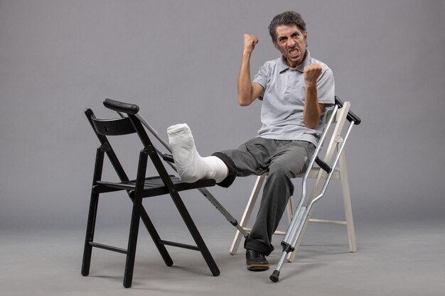 Vue de face jeune homme assis avec pied cassé et béquilles sur mur gris douleur au pied accident jambe cassée torsion