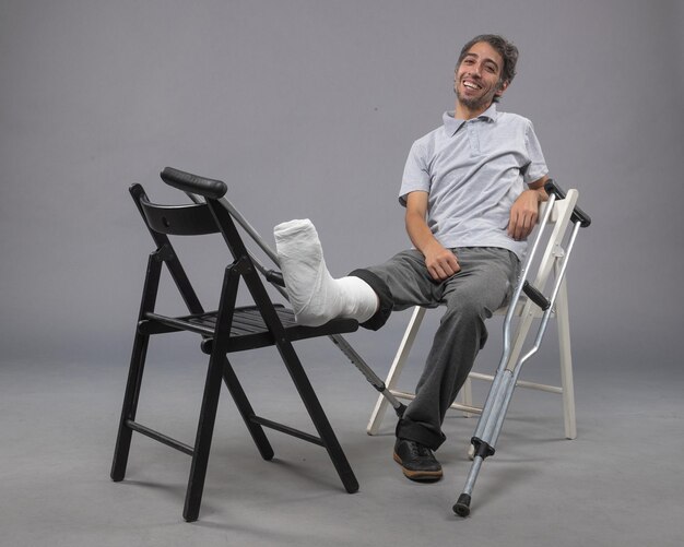 Vue de face jeune homme assis avec pied cassé et béquilles se sentant heureux sur un mur gris accident torsion douleur jambe cassée