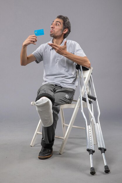 Vue de face jeune homme assis avec pied cassé tenant une carte bancaire bleue sur le mur gris douleur pied cassé accident torsion mâle