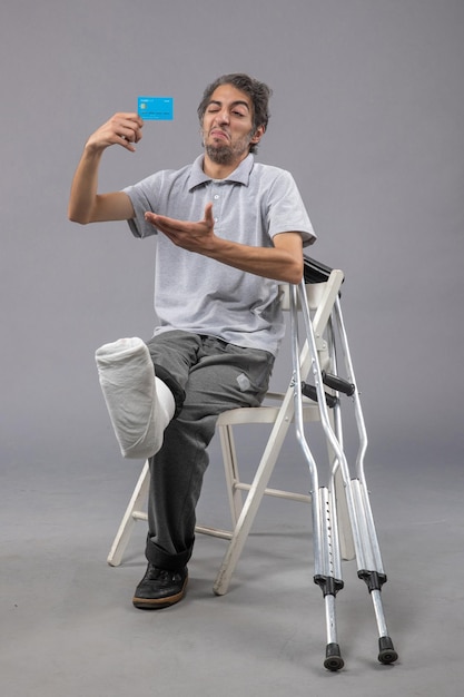 Vue de face jeune homme assis avec pied cassé tenant une carte bancaire bleue sur sol gris accident cassé douleur au pied torsion mâle