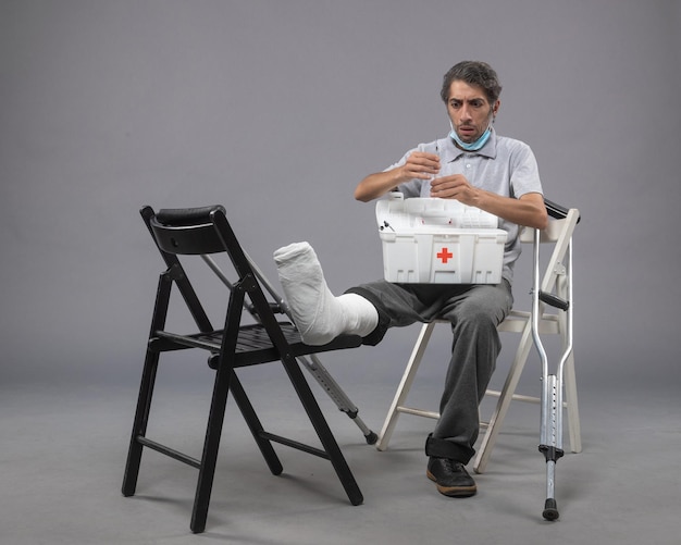 Vue de face jeune homme assis avec un pied cassé et tenant une injection sur un mur gris médecine torsion mâle douleur pieds jambes cassées