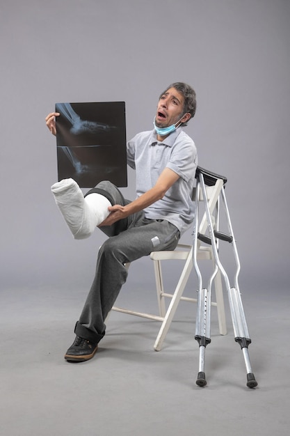 Vue de face jeune homme assis avec un pied cassé et tenant une radiographie de celui-ci sur le mur gris douleur jambe accident pied torsion mâle