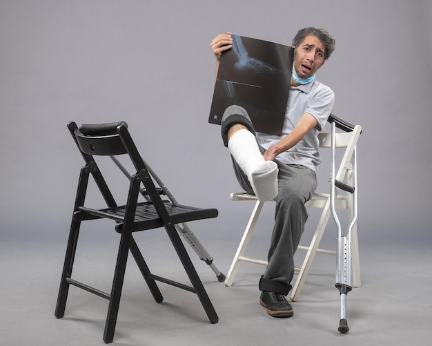 Vue de face jeune homme assis avec un pied cassé et tenant une radiographie de celui-ci sur un mur gris douleur torsion jambes cassé pied accidenté masculin