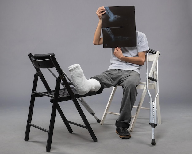 Vue de face jeune homme assis avec un pied cassé et tenant une radiographie de celui-ci sur le mur gris torsion douleur jambe cassée pied accidenté masculin
