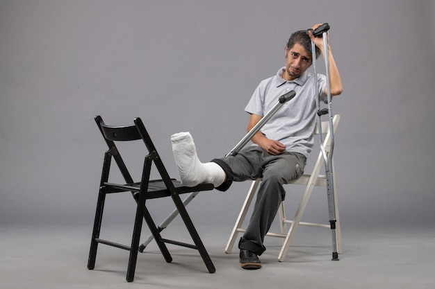 Vue de face jeune homme assis avec pied cassé et utilisant des béquilles sur le mur gris pied torsion douleur accident jambes cassées