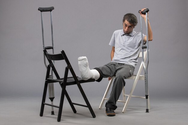 Vue de face jeune homme assis avec pied cassé et utilisant des béquilles sur le mur gris pied torsion douleur cassée accident jambes