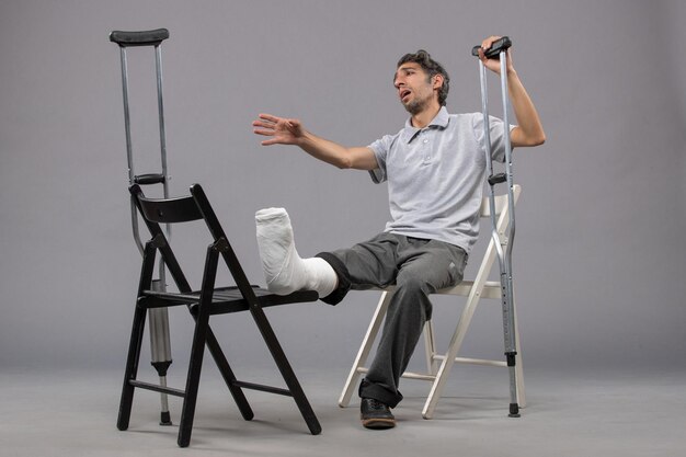 Vue de face jeune homme assis avec un pied cassé et utilisant des béquilles sur un mur gris torsion cassée douleur pied accident jambe