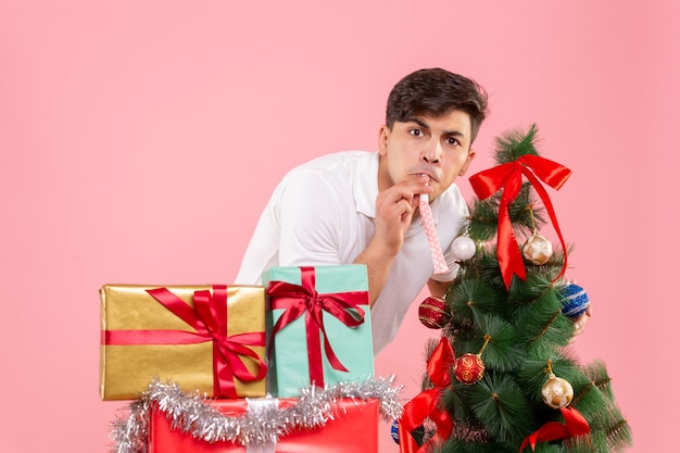 Vue de face jeune homme autour des cadeaux de Noël et arbre de vacances sur le fond rose