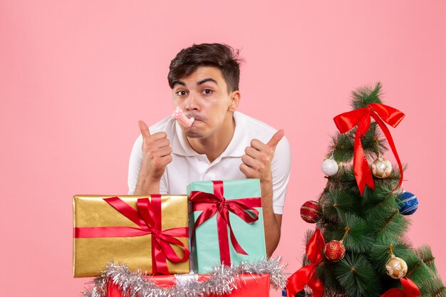 Vue de face jeune homme autour des cadeaux de Noël et arbre de vacances sur le fond rose