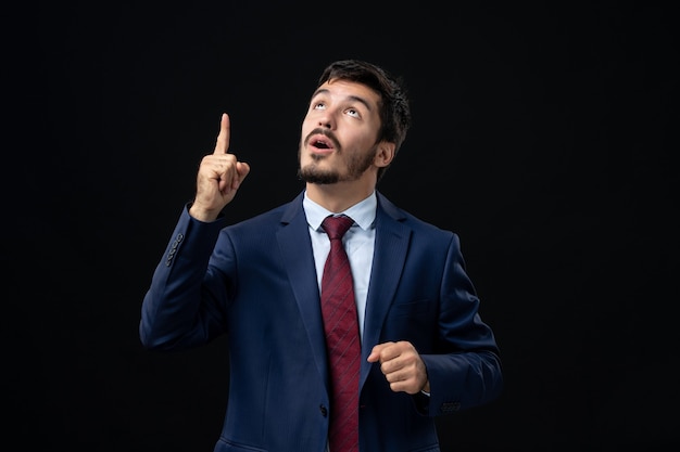 Vue de face d'un jeune homme barbu confus et émotif en costume pointant vers le haut sur un mur sombre isolé