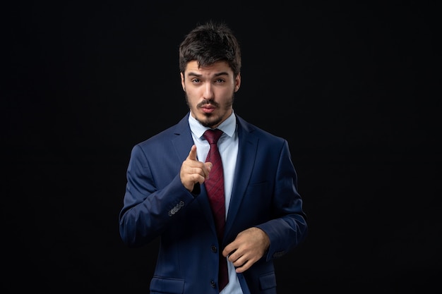 Vue de face d'un jeune homme barbu insatisfait pointant quelque chose vers l'avant sur un mur sombre isolé