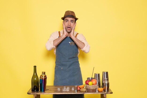 Vue de face jeune homme barman devant la table avec des boissons shakers sur un bureau jaune