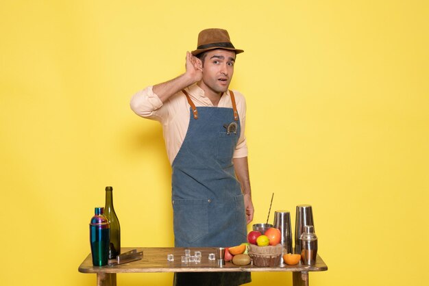 Vue de face jeune homme barman devant la table avec des boissons shakers sur un bureau jaune