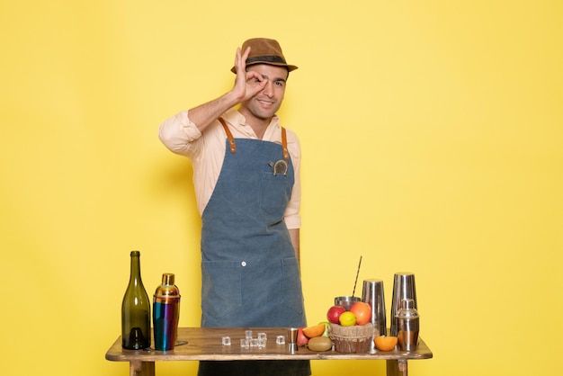 Vue de face jeune homme barman devant table avec shakers et boissons souriant sur fond jaune