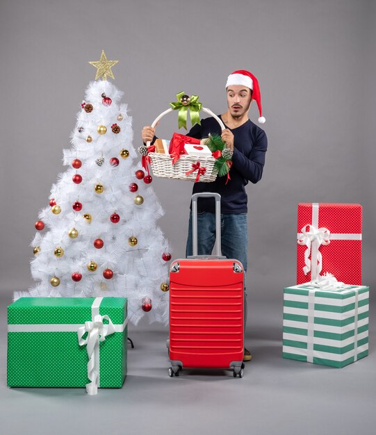 Vue de face jeune homme avec bonnet de noel regardant panier-cadeau près de l'arbre de Noël sur isolé