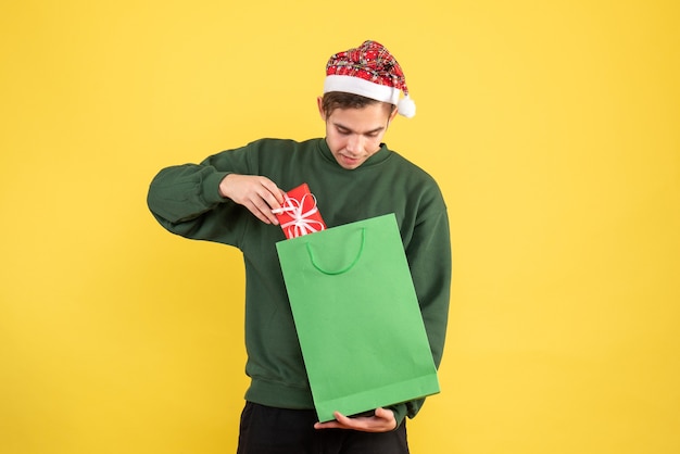 Vue de face jeune homme avec Bonnet de Noel tenant un sac à provisions vert et cadeau regardant cadeau sur fond jaune