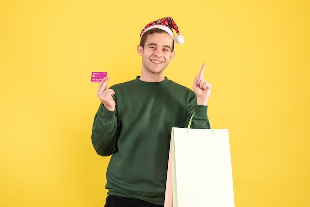 Vue de face jeune homme avec bonnet de Noel tenant des sacs à provisions et carte debout sur fond jaune copie espace