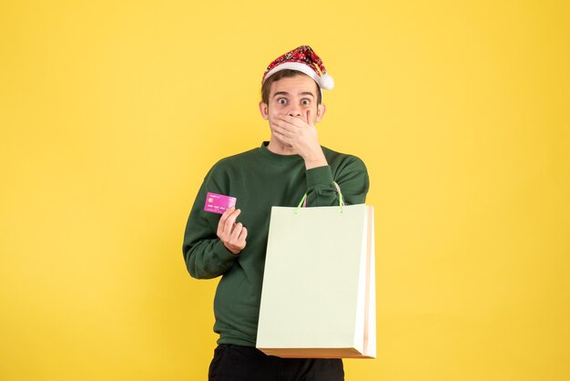 Vue de face jeune homme avec bonnet de Noel tenant des sacs à provisions et carte mettant la main à sa bouche sur fond jaune