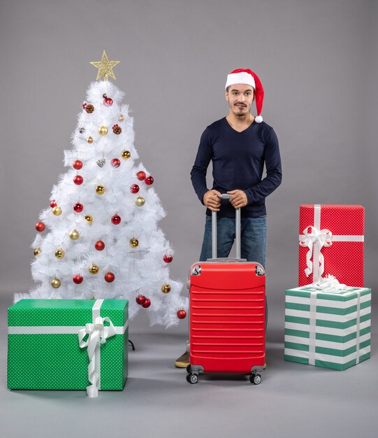 Vue de face jeune homme avec bonnet de noel tenant une valise debout près de l'arbre de Noël sur isolé