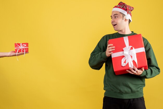 Vue de face jeune homme avec cadeau de Noël en regardant le cadeau en main féminine sur fond jaune