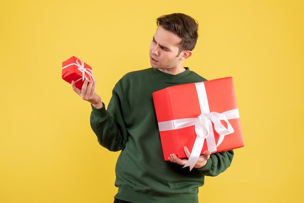 Vue de face jeune homme avec cadeau de Noël vérifier son cadeau debout sur fond jaune