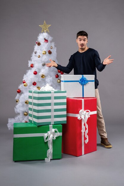 Vue de face jeune homme avec des cadeaux et arbre de noël sur un bureau gris cadeau noël nouvel an