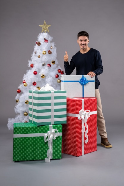 Vue de face jeune homme avec des cadeaux et arbre de Noël sur fond gris