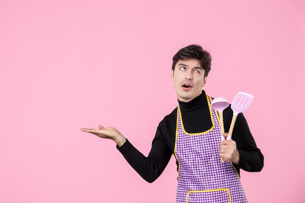 Vue de face jeune homme en cape tenant des cuillères roses sur fond rose profession cuisine travail cuisine repas pâte couleur uniforme horizontale