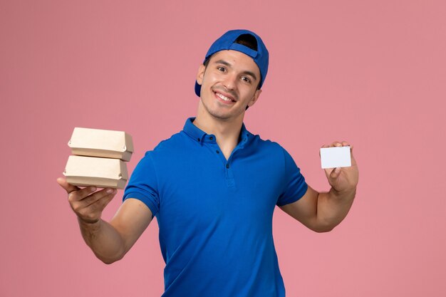 Vue de face jeune homme en cape uniforme bleu tenant peu de colis alimentaires de livraison avec carte sur mur rose clair