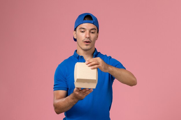 Vue de face jeune homme en cape uniforme bleu tenant peu de colis alimentaires de livraison sur le mur rose