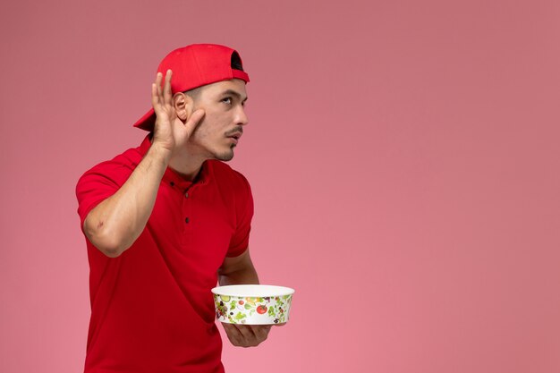 Vue de face jeune homme en cape uniforme rouge tenant le bol de livraison et en essayant d'entendre sur fond rose clair.