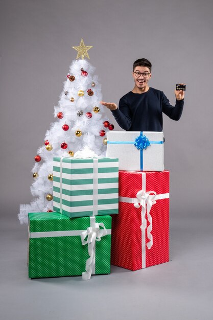 Vue de face jeune homme avec carte bancaire sur un bureau gris présente noël nouvel an