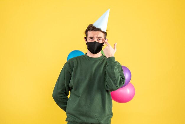 Vue de face jeune homme avec chapeau de fête et ballons colorés debout sur fond jaune