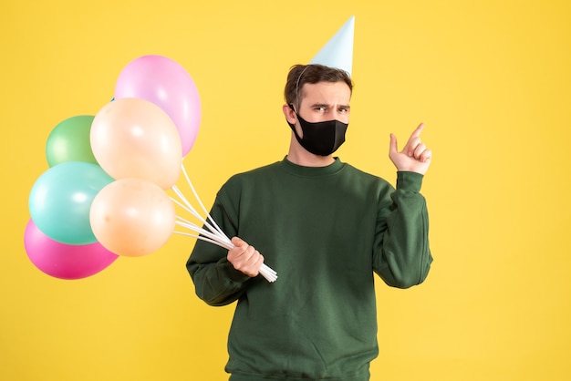 Vue de face jeune homme avec chapeau de fête et ballons colorés pointant vers l'arrière debout sur jaune