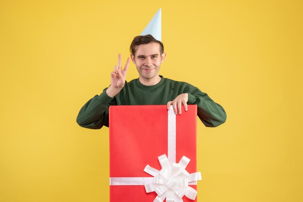 Vue de face jeune homme avec chapeau de fête faisant signe de la victoire debout derrière une grande boîte-cadeau sur fond jaune