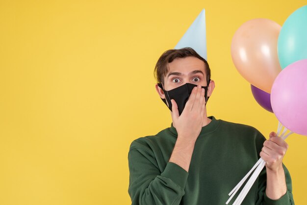 Vue de face jeune homme avec chapeau de fête et masque noir tenant des ballons sur jaune