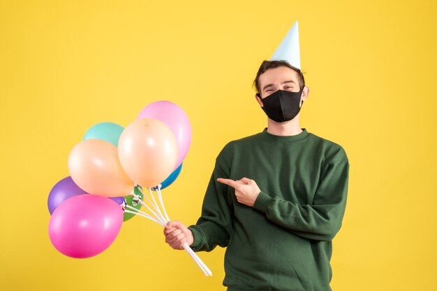 Vue de face jeune homme avec chapeau de fête pointant sur des ballons colorés debout sur fond jaune