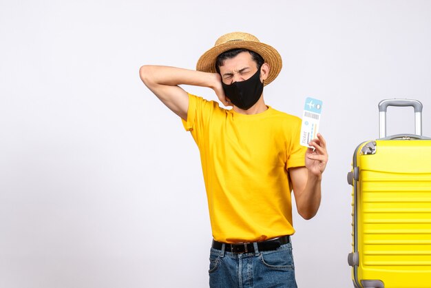 Vue de face jeune homme avec chapeau de paille debout près de valise jaune tenant un billet de voyage et son oreille